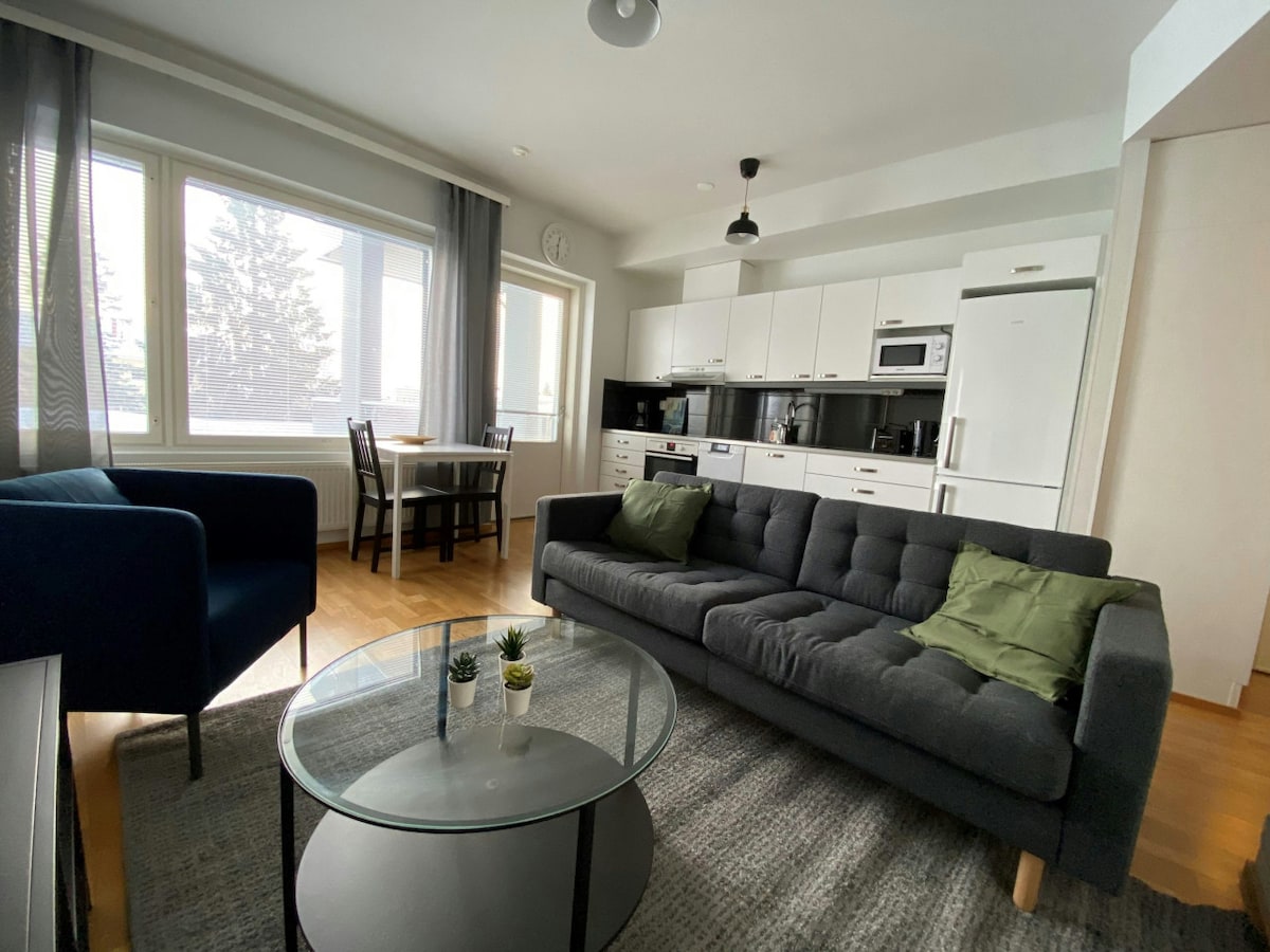 The living area showcases a comfortable sofa with grey upholstery, accompanied by two green cushions. A circular glass table sits atop a soft area rug. The adjoining kitchen is equipped with modern appliances, while large windows allow natural light to illuminate the space.