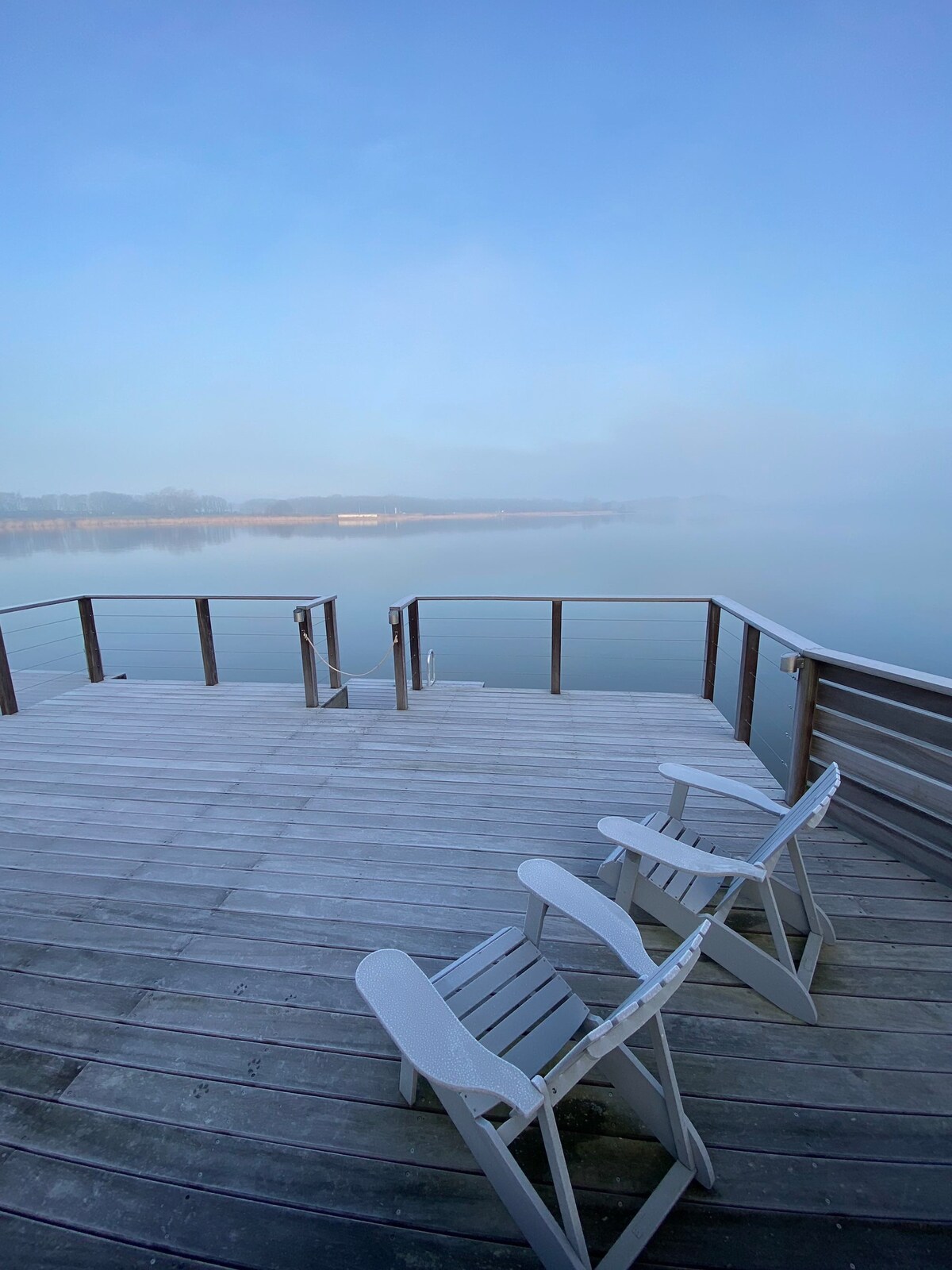 A wooden deck extends over calm water, featuring two white deck chairs facing towards the serene landscape. Soft fog blankets the horizon, creating a tranquil atmosphere. The railing provides an unobstruck view of the peaceful surroundings.