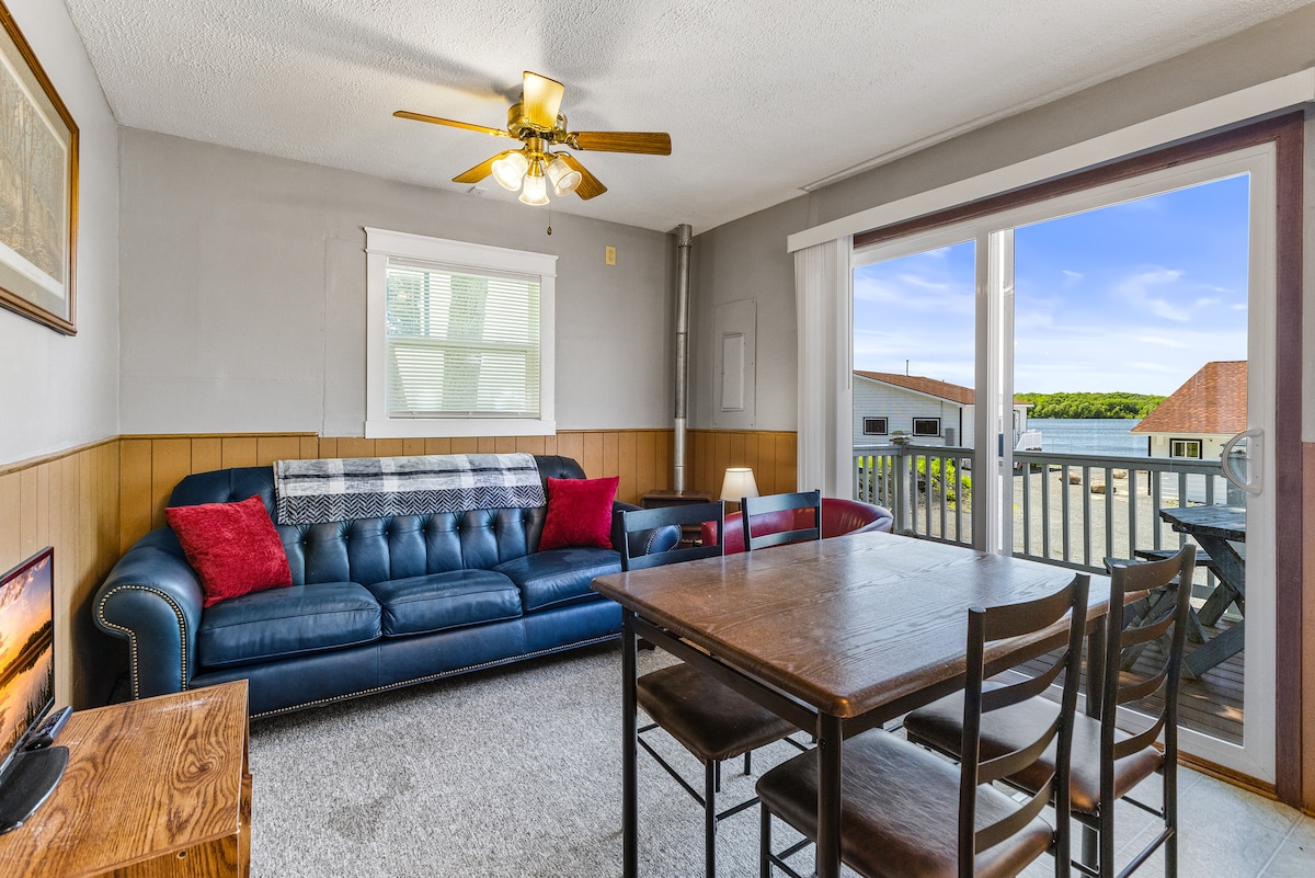 A spacious living area features a blue sectional sofa with red accent pillows, complemented by a wooden dining table with four chairs. Natural light floods the room through sliding glass doors, which provide views of the outdoor deck and surrounding area.