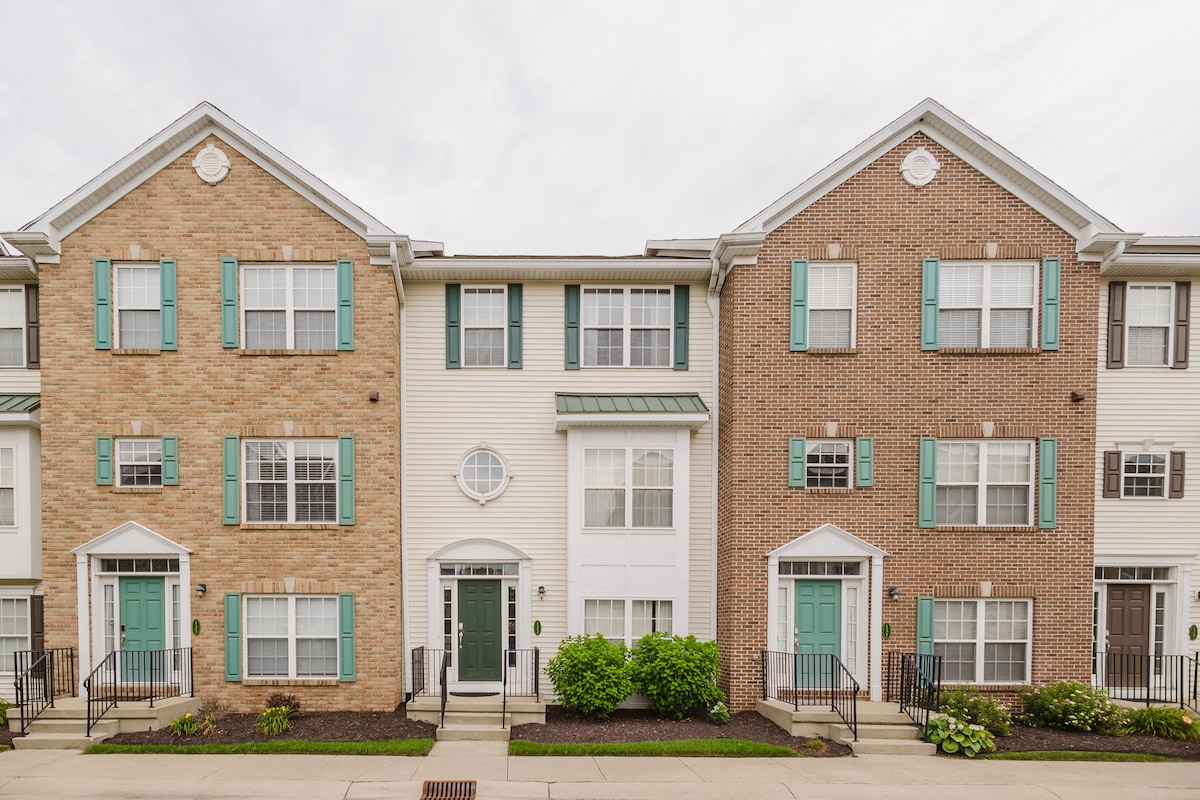 A row of townhomes showcases a blend of brick and siding exteriors. Each unit features a decorative entrance with a green door and white trim. Windows are evenly spaced along the facade, framed by greenery and well-maintained landscaping, providing a welcoming street view.