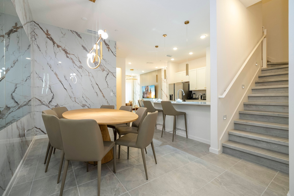 A modern dining area is highlighted by a round wooden table surrounded by six upholstered chairs. A marble accent wall adds a touch of elegance, while pendant lighting enhances the open-concept layout leading to a fully equipped kitchen and staircase.
