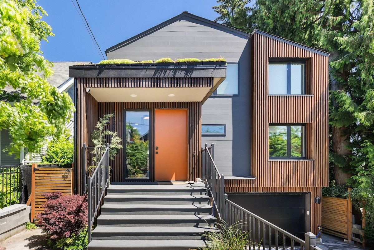 The modern home features a striking orange front door and a combination of natural wood accents and large windows. Stairs lead up to the entrance, framed by greenery and landscaping that enhance the overall appeal of the exterior.