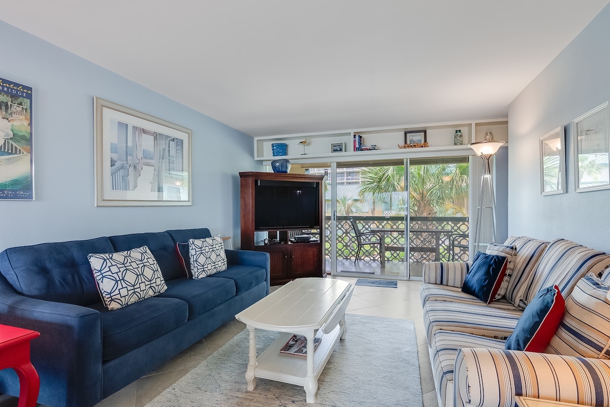 A comfortably arranged living area features two sofas, one in a deep blue hue and another with stripes. A light coffee table is centered between them. A TV unit is visible with a large screen. Natural light enters through sliding glass doors leading to a balcony.