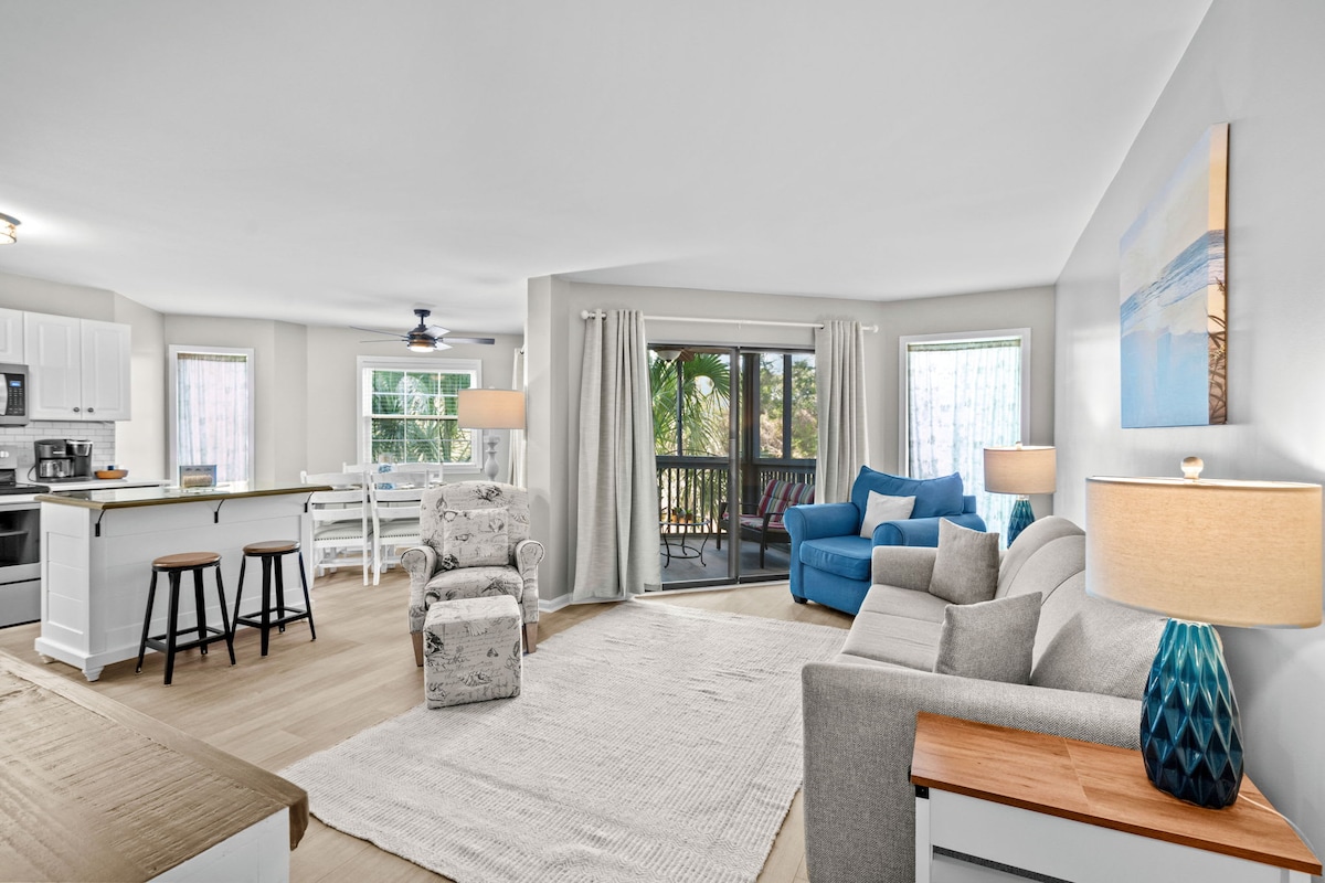 A spacious living area features a comfortable seating arrangement with a light-colored sofa, blue accent chair, and a recliner. Natural light fills the room through large windows with sheer curtains, and a dining area with white chairs complements the open layout. A balcony can be seen through the glass doors.