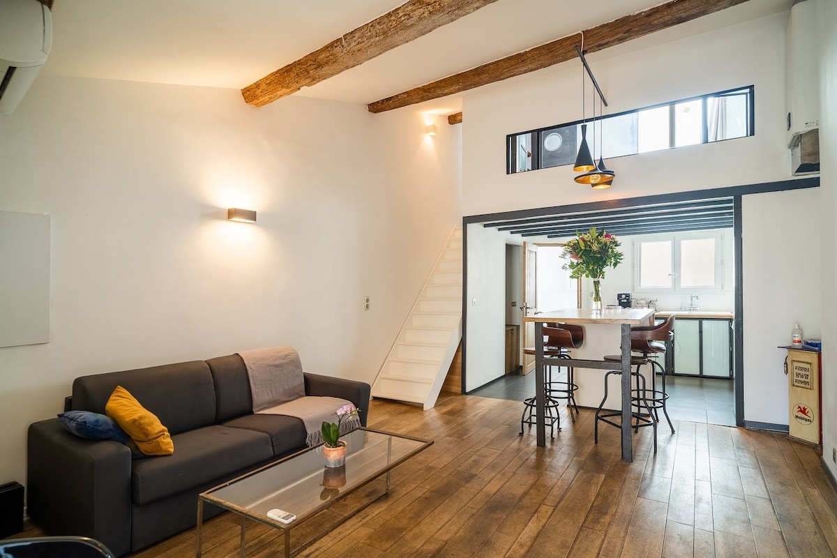 An inviting living area features a dark sofa with a colorful accent pillow and a glass coffee table. Above, wooden beams enhance the space's character. The dining area includes tall bar stools around a white counter. A staircase leads to another level, with a glimpse of a bright kitchen.