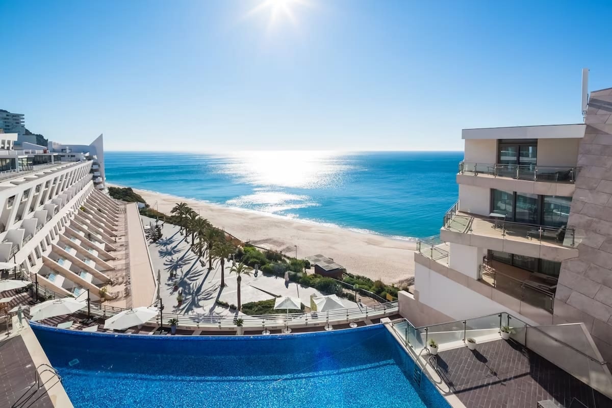 A scenic view of the outdoor pool is displayed, with its water reflecting the sunlight. Adjacent to the pool, sun loungers are arranged along the beach, which stretches out alongside the shimmering ocean. The clear blue sky completes this tranquil coastal setting.