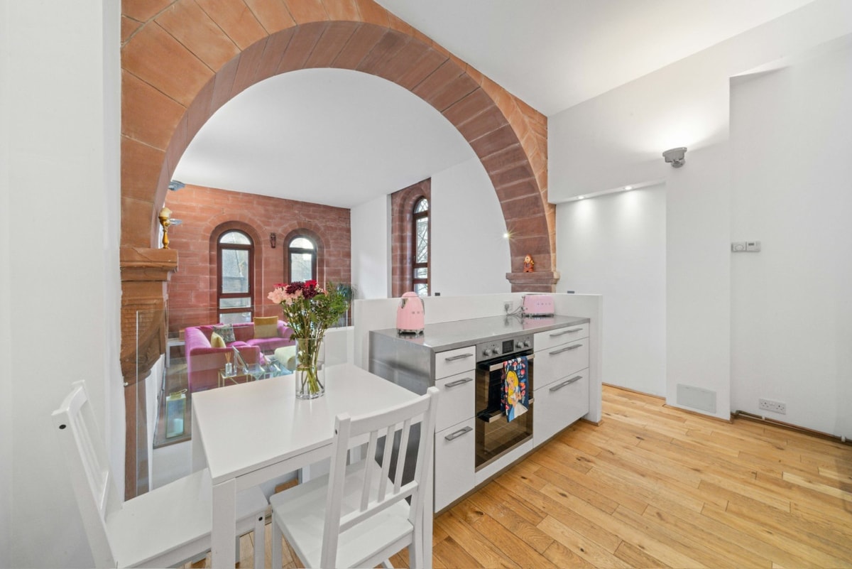 The open-plan kitchen features modern appliances with a sleek countertop and kitchen island. A small dining table with white chairs is positioned nearby. Archway details lead into a bright living area with large arched windows, enhancing the spaciousness of the design.