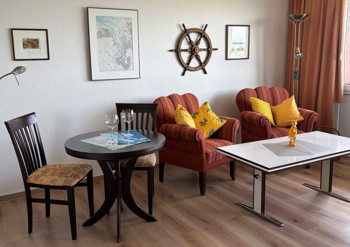 A living area is presented with two armchairs featuring yellow cushions and a nautical decor element on the wall. A round dining table is accompanied by a single chair, while large windows provide natural light, highlighting the warm wooden flooring and textured wall.