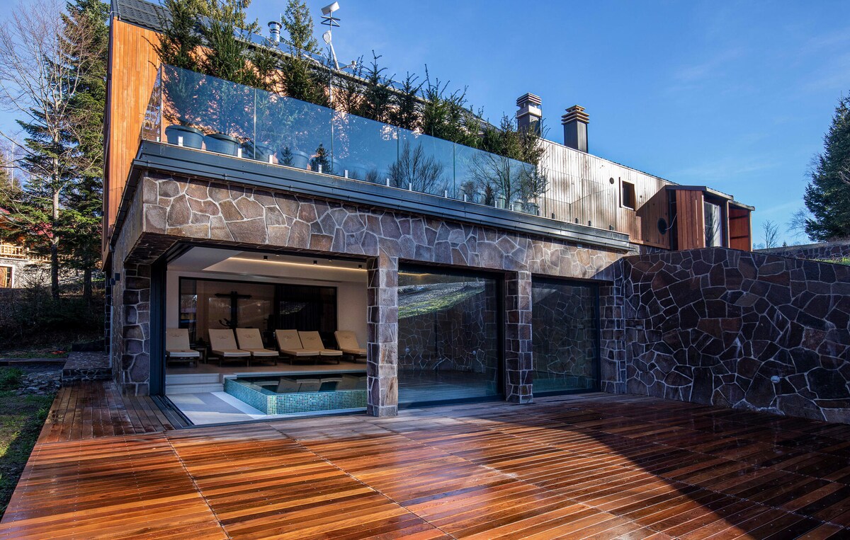 An elegant outdoor area features a polished wooden deck that extends towards a glass-enclosed living space. A relaxing whirlpool is visible beside sun loungers. The backdrop includes a stone-clad wall and a modern structure, surrounded by greenery and sunlight.
