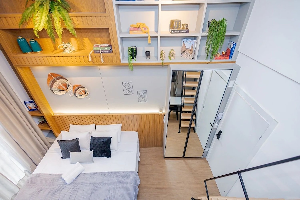 An overhead view of a welcoming bedroom is displayed, featuring a spacious bed with crisp white linens and decorative pillows. A mirrored wardrobe reflects the surroundings. The walls are lined with wooden shelving, adorned with plants and decorative items, enhancing the room's aesthetic.