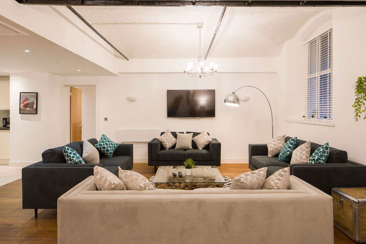 A spacious living room features ample seating with two sofas and several cushioned chairs. A minimalist coffee table sits in the center, complemented by decorative pillows on the sofas. A wall-mounted TV and a stylish floor lamp complete the inviting space.