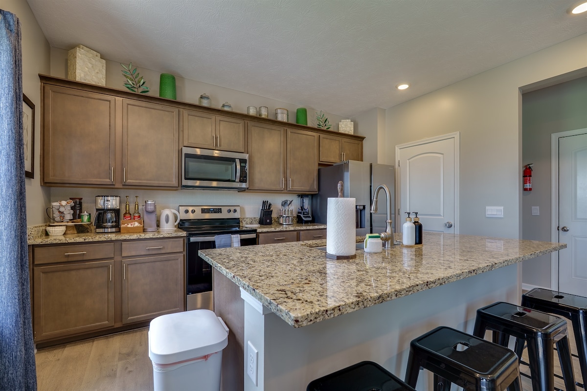 The kitchen features granite countertops with a spacious island equipped with seating for four. Dark wooden cabinets hold various kitchen essentials. Stainless steel appliances, including a microwave and oven, complement the space. Light streams in from a nearby window, adding brightness to the decor.