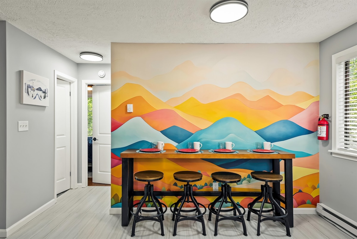 A dining area showcases a vibrant mural depicting rolling hills in warm colors. A wooden table with four black stools is set for breakfast, featuring white mugs and red plates. Natural light filters in through the window, enhancing the cheerful atmosphere.