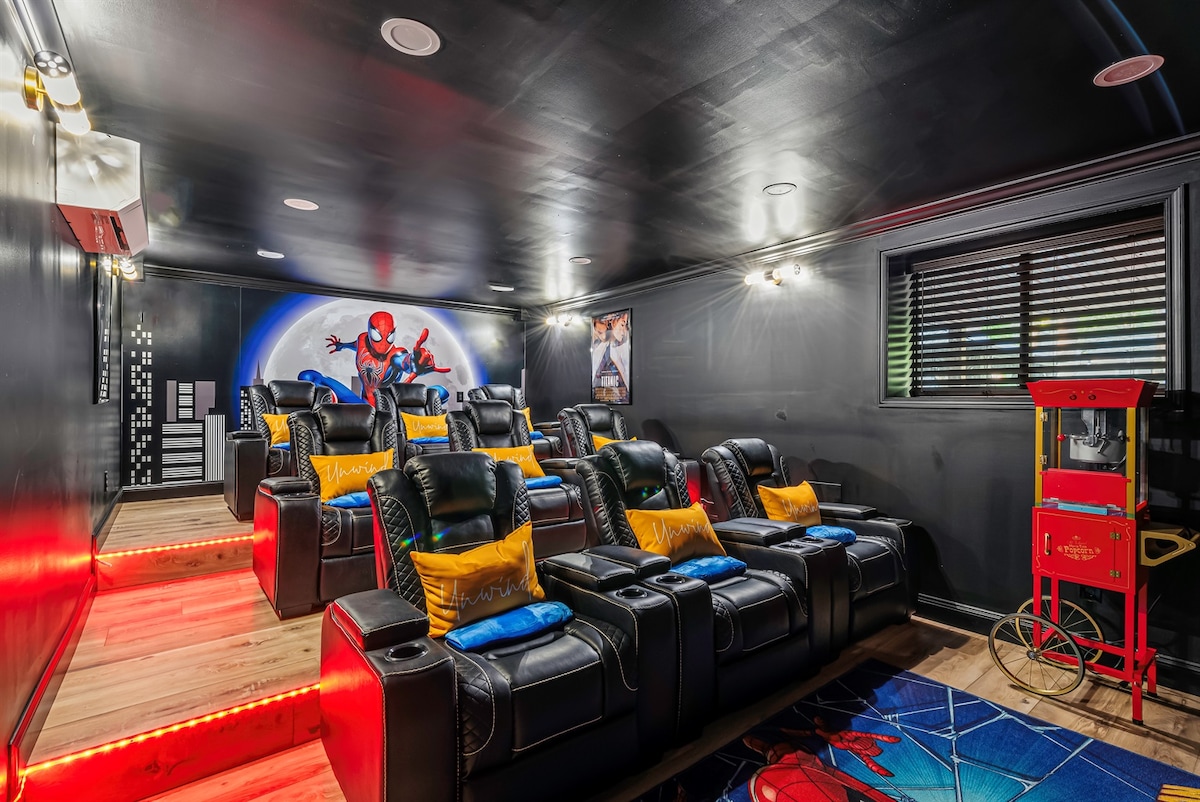 The home theater features comfortable reclining seats arranged in tiers, illuminated by ambient lighting. A vibrant mural of a superhero decorates the wall, and a popcorn machine stands near the entrance, adding to the cinematic experience.