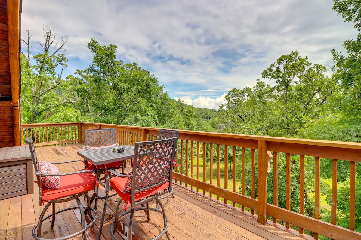 A spacious outdoor deck is furnished with a round dining table and four chairs, complemented by colorful cushions. The deck overlooks a lush green landscape filled with trees, creating a peaceful and scenic setting.