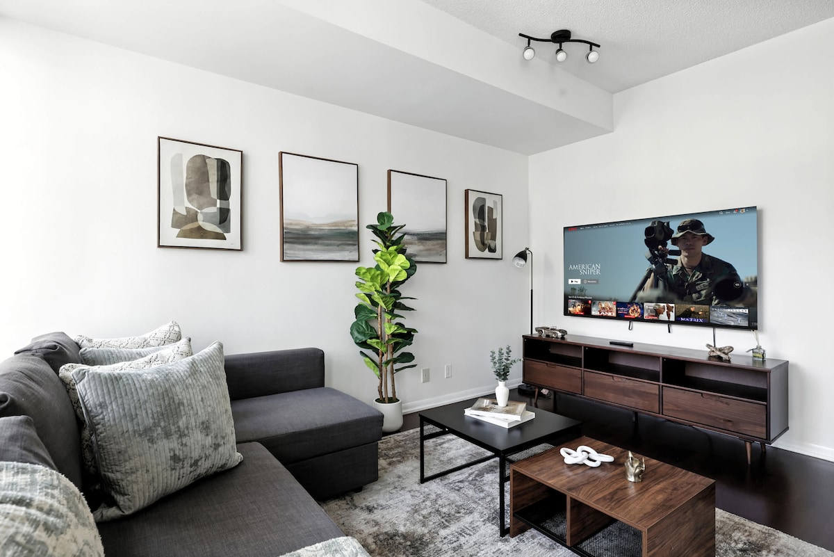 The living area features a modern sectional sofa adorned with plush cushions, complemented by a sleek coffee table. A large smart TV is mounted on a media console, with framed artwork arranged on the wall. A potted plant adds a touch of greenery, enhancing the ambiance.