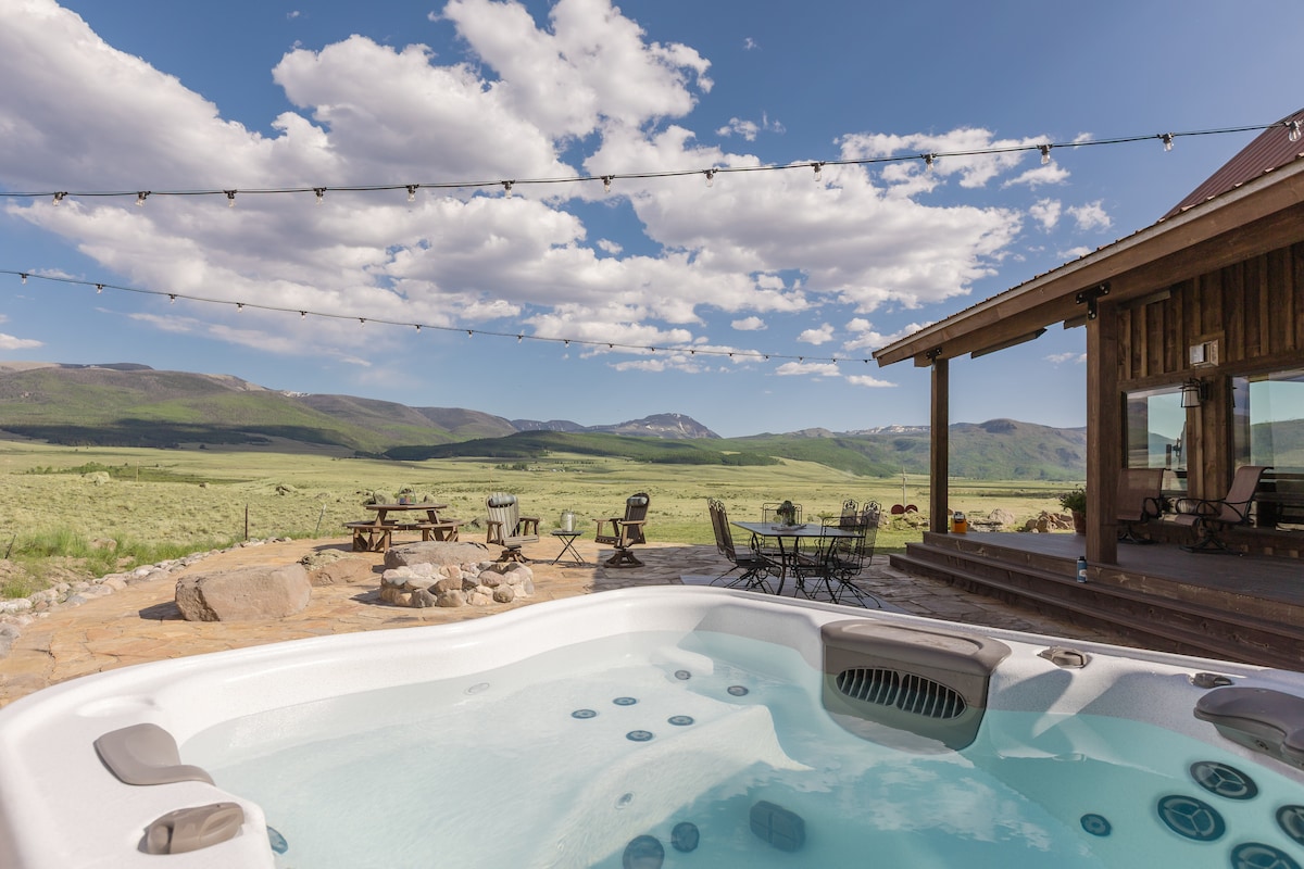 A hot tub is positioned on a stone patio, surrounded by natural landscaping and offering expansive views of rolling hills and mountains. String lights are draped overhead, adding a warm touch. Nearby, a seating area with tables and chairs invites relaxation amid the serene environment.