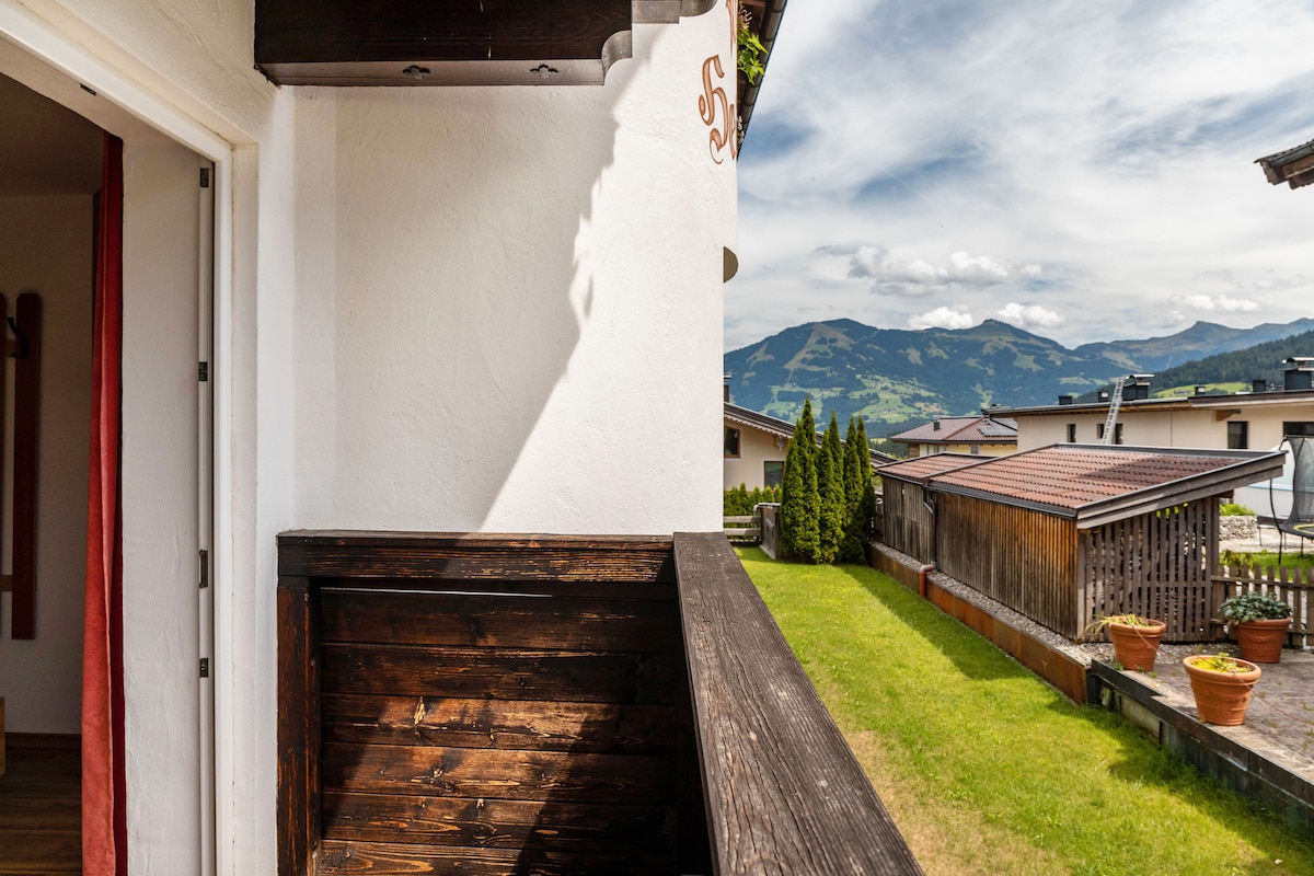 A private balcony offers scenic views of the surrounding mountains and lush valley below. Wood accents create a natural and inviting space, while the sunlight casts gentle shadows on the balcony railing. Green grass and potted plants are visible in the foreground.