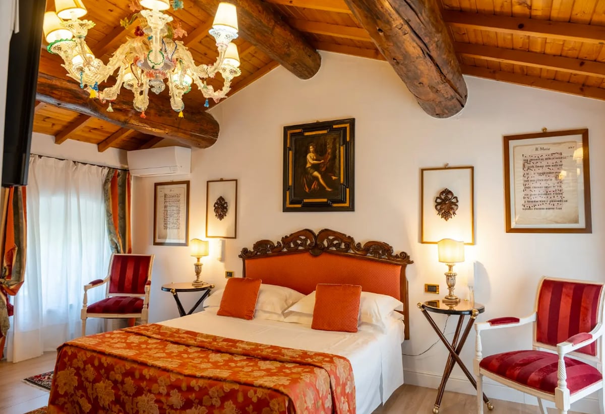 A spacious bedroom features a large bed adorned with an elegant floral coverlet. Two armchairs are placed beside a bedside table, and decorative wall art complements the room's warm wooden beams and ceiling. Soft light from lamps enhances the cozy ambiance.