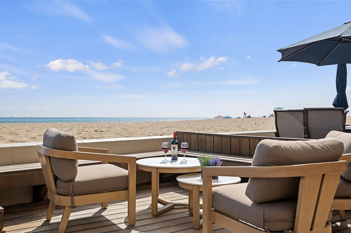 A private beachfront patio is showcased, featuring comfortable lounge chairs paired with a round table. Glasses and a bottle are neatly arranged on the table, with soft sand and a calm ocean view in the background under a clear blue sky.