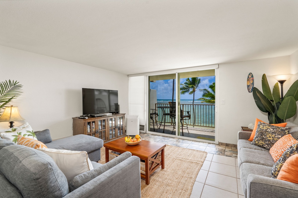 A welcoming living area features comfortable seating arrangements, inclusive of a gray sectional and a light beige couch adorned with colorful cushions. A wooden coffee table rests between them. Large sliding glass doors lead to a private lanai, offering views of the ocean and swaying palm trees.