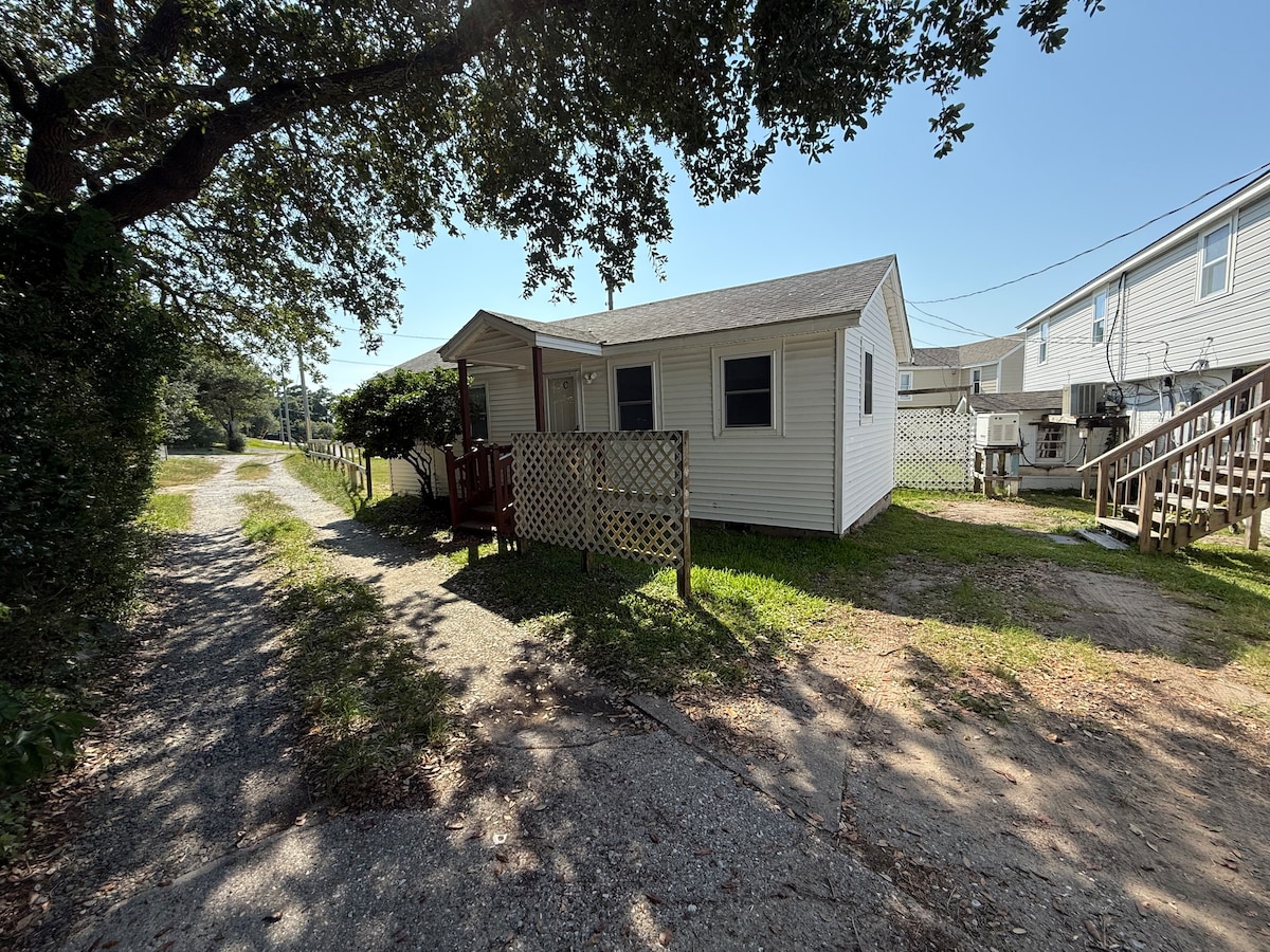 The exterior of the cottage features a simple white façade and is framed by greenery. A gravel path leads to the entrance, which is complemented by wooden railing and lattice privacy panels. Nearby, two staircases provide access to separate levels of surrounding structures.