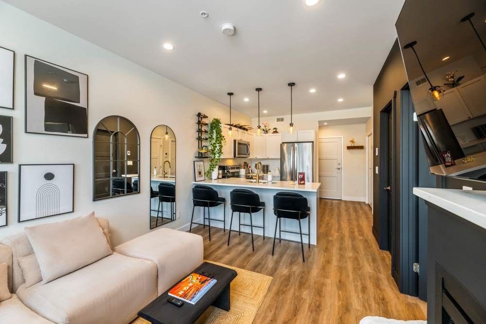 A modern living area is showcased with light wood flooring and a comfortable sectional sofa. A sleek kitchen bar with black stools and pendant lighting complements the space. Wall art and mirrors enhance the aesthetic, while a small table adds a functional touch.