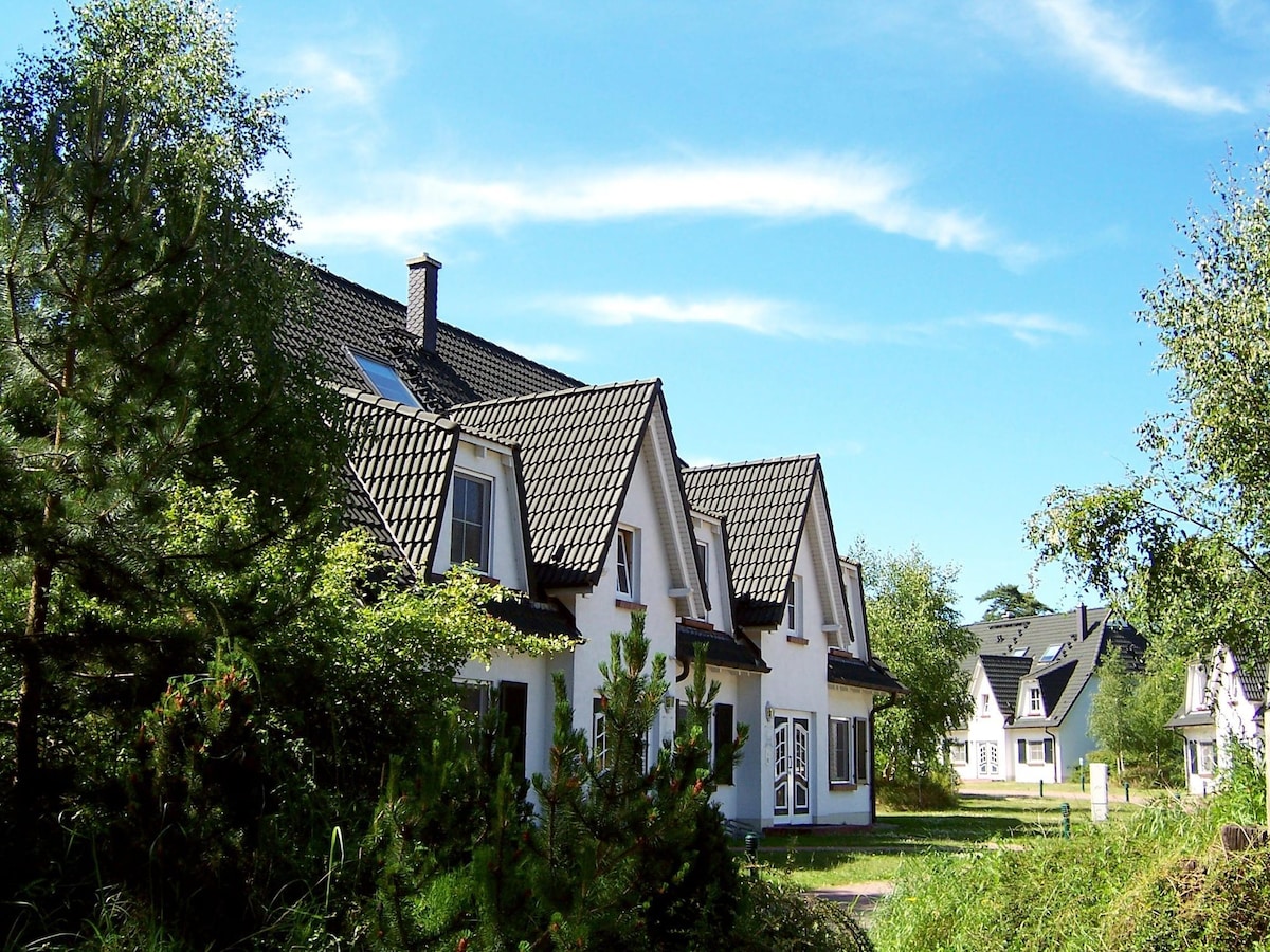 The accommodation features a two-story structure with a tiled roof, surrounded by greenery. Large windows dominate the front, allowing natural light to fill the interior. Nearby, additional similar buildings are visible, set against a clear blue sky.