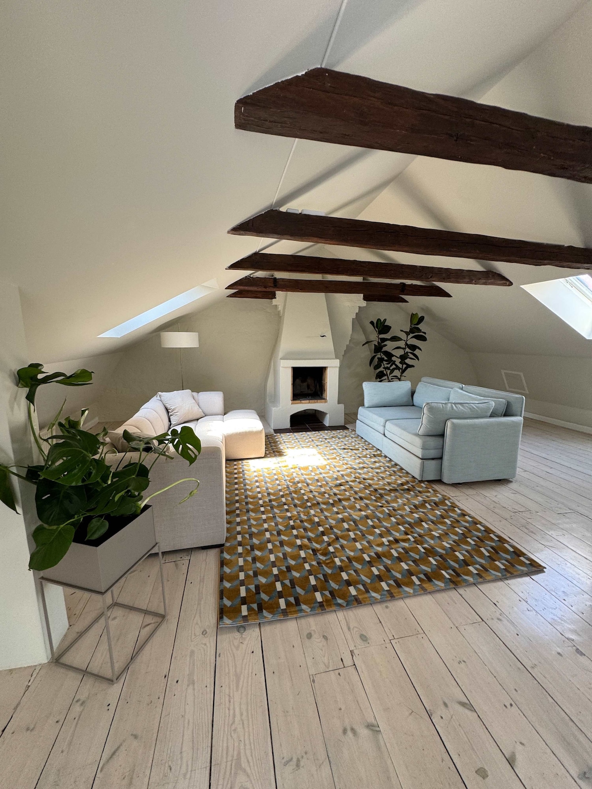 The spacious living room features exposed wooden beams, enhancing the room's character. A cozy sectional sofa and a light blue sofa are arranged on a patterned area rug. A potted plant adds a touch of greenery, and natural light streams in through skylights.