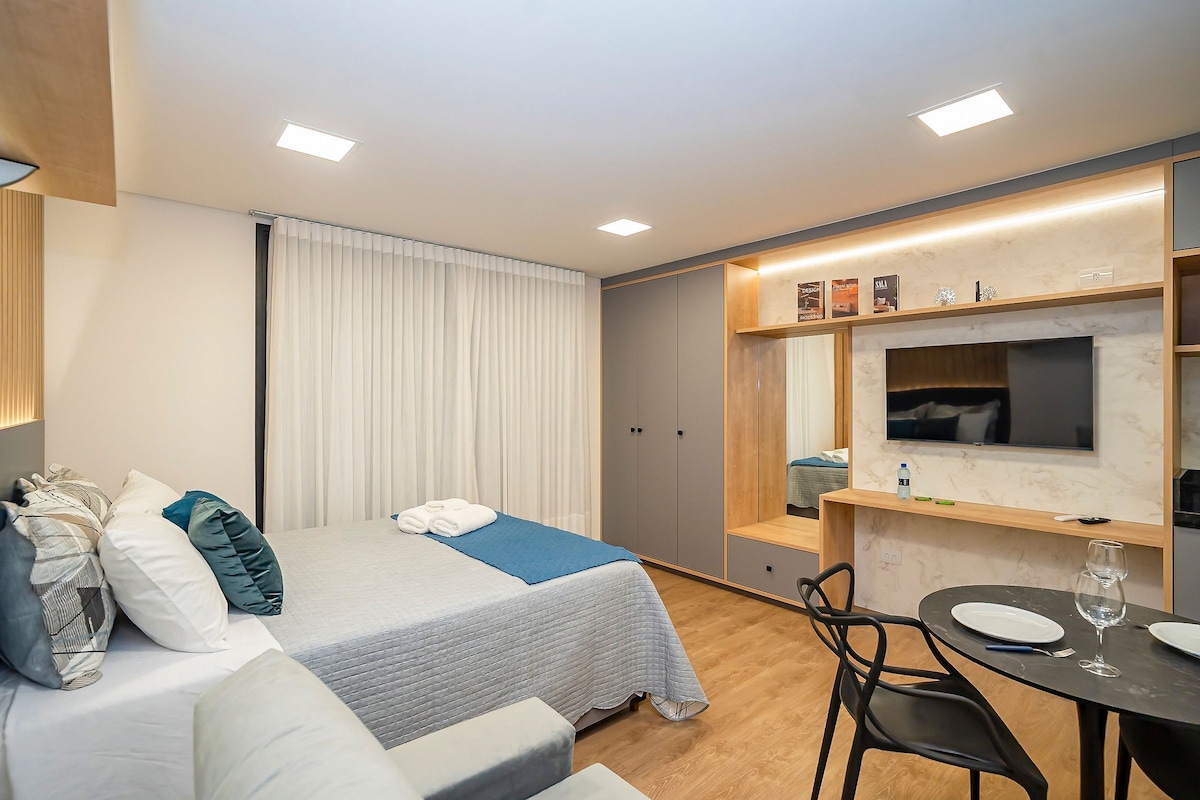A modern studio apartment is showcased, featuring a spacious bed with decorative pillows. A cozy seating area is positioned beside a small dining table, while a wall-mounted television and a mirrored cabinet add functionality. Soft, natural light filters through sheer curtains, enhancing the muted color palette.