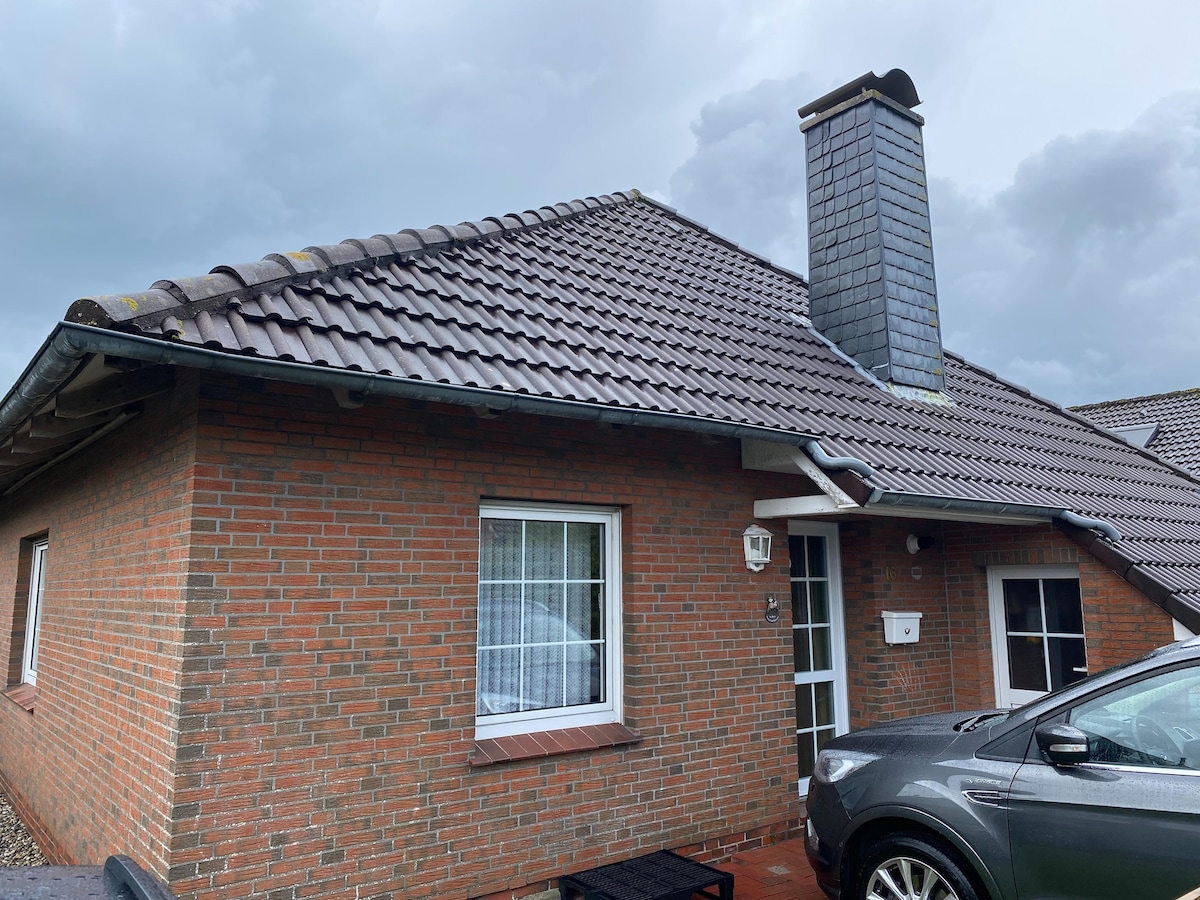 The exterior of the semi-detached holiday home is shown, featuring a brick facade and a sloped tiled roof. A small porch with a door is visible, along with a window adorned with white curtains. A parked vehicle is present in the foreground.