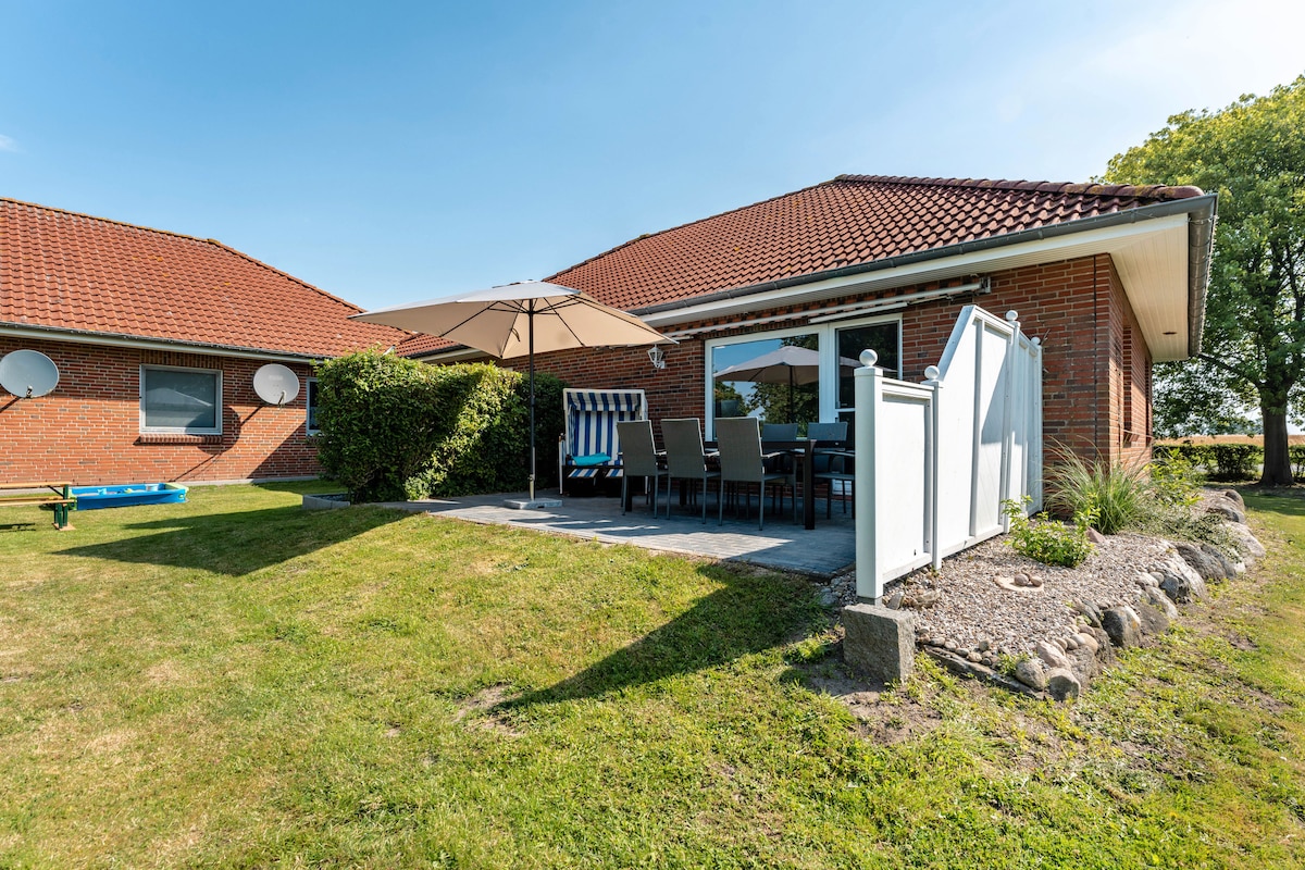 An outdoor terrace is presented, featuring a large umbrella shading a dining table surrounded by several chairs. The well-maintained garden is visible, with greenery and a stone border surrounding the patio area. Two satellite dishes are mounted on the nearby wall.