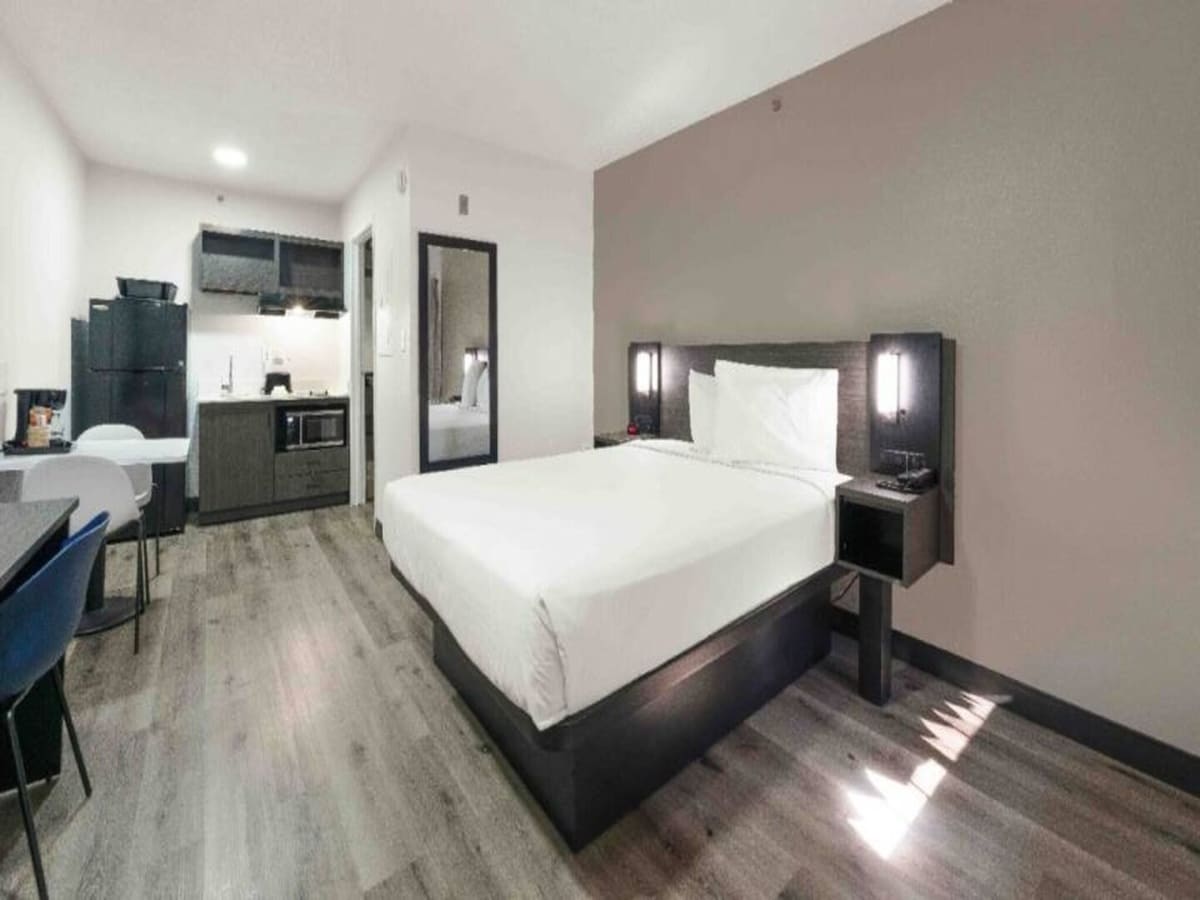 A studio room features a spacious bed with a nightstand on one side. A small kitchenette with modern appliances is visible, along with a dining table and chair. The room has wood-like flooring and neutral wall colors, creating a clean and comfortable environment.