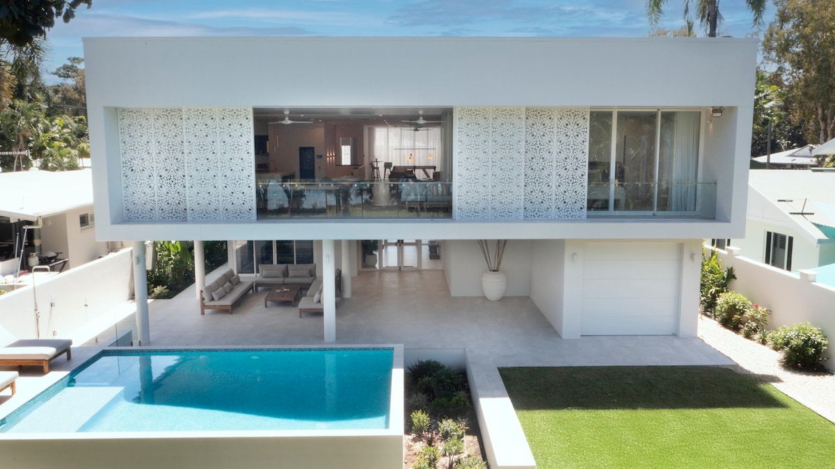 The exterior view highlights a contemporary two-story home with intricate decorative panels. A spacious terrace features a rectangular pool and a lounge area. Lush greenery surrounds the property, enhancing the coastal ambiance. Large glass doors allow natural light to fill the interior space.