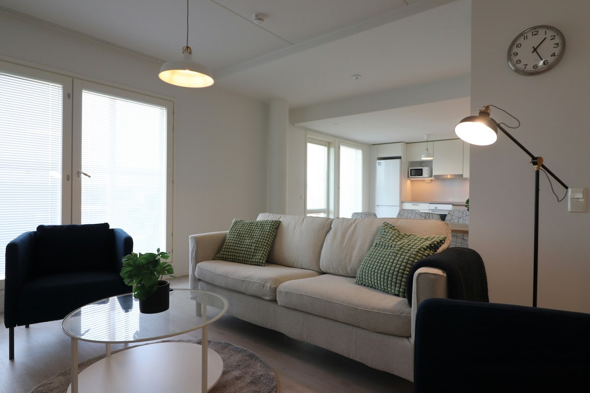 A modern living area is presented with a comfortable sofa and two armchairs. Natural light filters through large windows, illuminating a glass coffee table with a small plant. The open-plan layout connects to a stylish kitchen in the background.