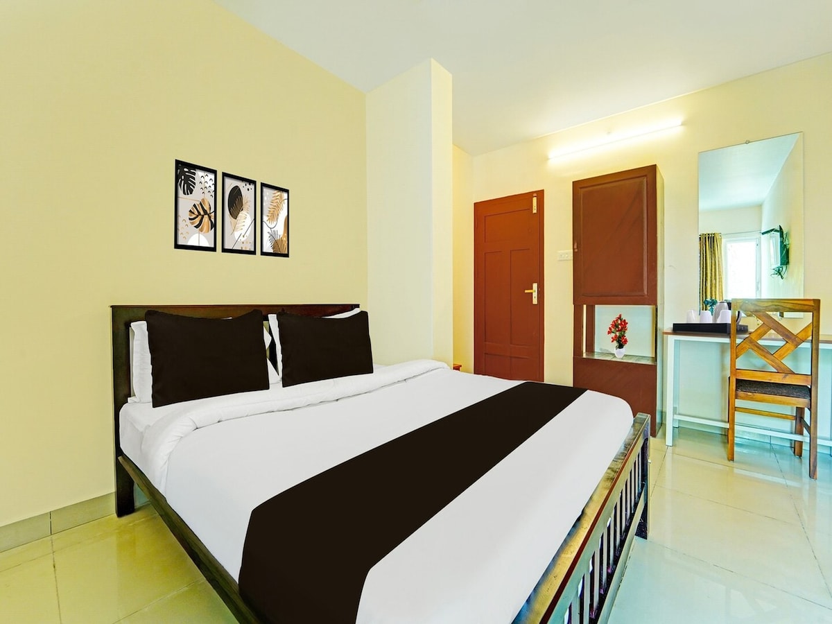 A spacious bedroom features a double bed adorned with a black and white bedspread. Accompanying decor includes three framed botanical prints on the wall. A wooden chair and table are situated beside a mirror, while natural light enters through a window dressed with light curtains.