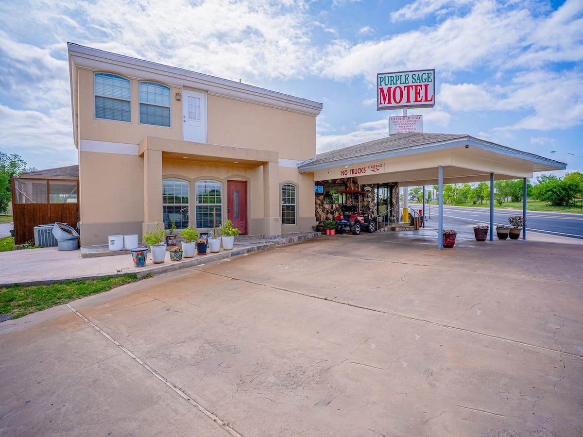 A two-story building is seen with a welcoming entrance and expansive front porch. Potted plants are arranged alongside the entrance. A prominent motel sign displays the name 'Purple Sage Motel.' Open parking space is available in front, with clear skies above.
