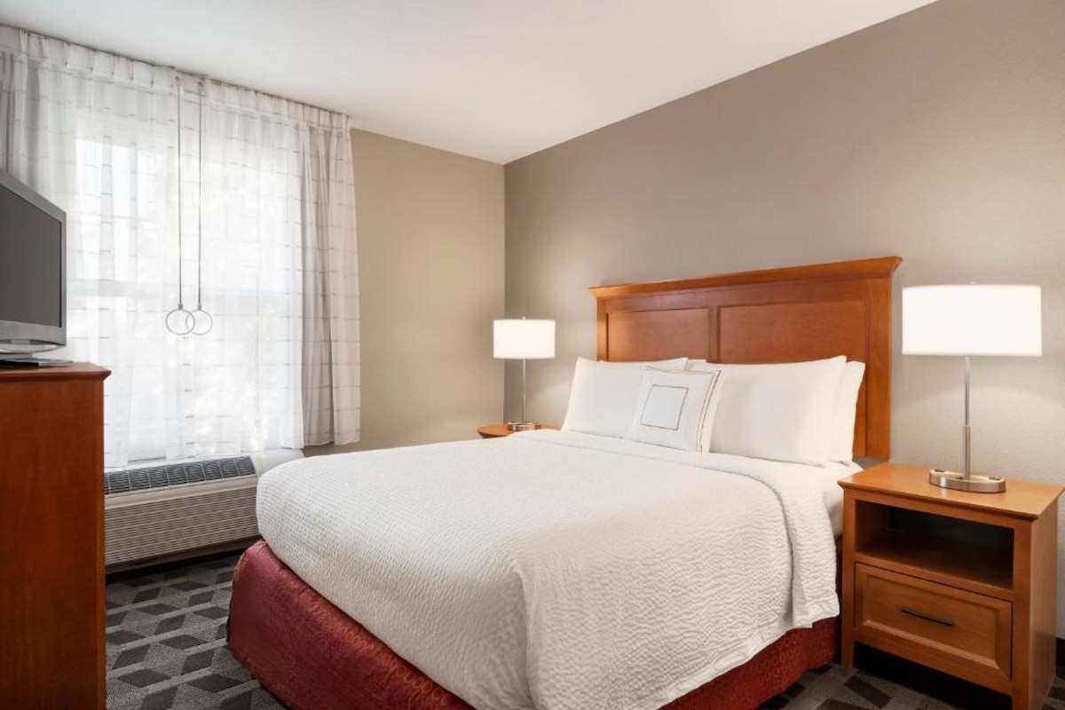 A spacious bedroom features a queen bed with a crisp white duvet. Two bedside lamps provide soft lighting. A flat-screen TV is mounted on a wooden dresser. Natural light floods in through a large window, adorned with sheer curtains.