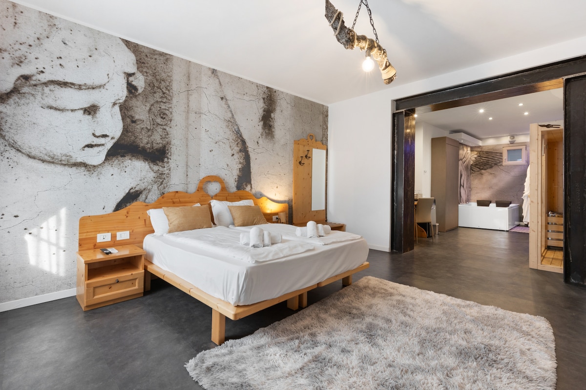 A spacious bedroom features a wooden bed with neatly arranged towels. A striking wall mural depicting a classical sculpture is contrasted by modern lighting. The room includes minimalist furniture and sleek flooring, with sunlight filtering through the window, enhancing the serene atmosphere.