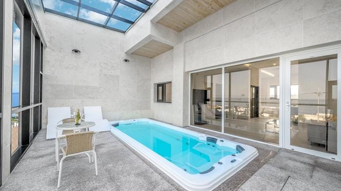 An outdoor terrace features a private pool with a clear blue surface, surrounded by stone walls. A small table and two chairs are positioned nearby. Large glass doors lead into an interior space filled with natural light from the skylights above.