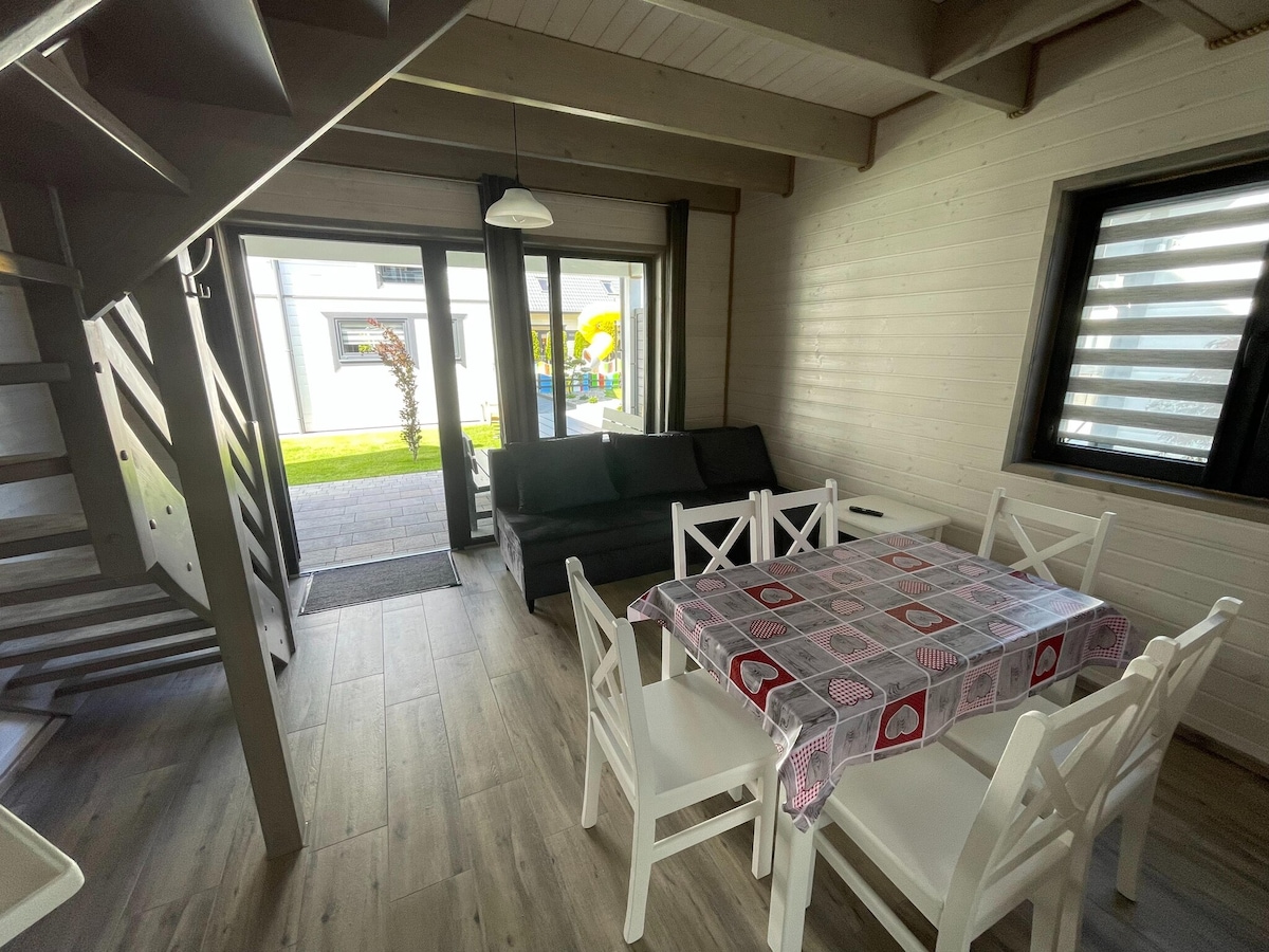 An open living area is seen featuring a dining table with six white chairs and a comfortable black sofa. Large glass doors provide access to the outdoor space, allowing natural light to fill the room. Wooden stairs lead to the upper level, while a pendant light hangs overhead.
