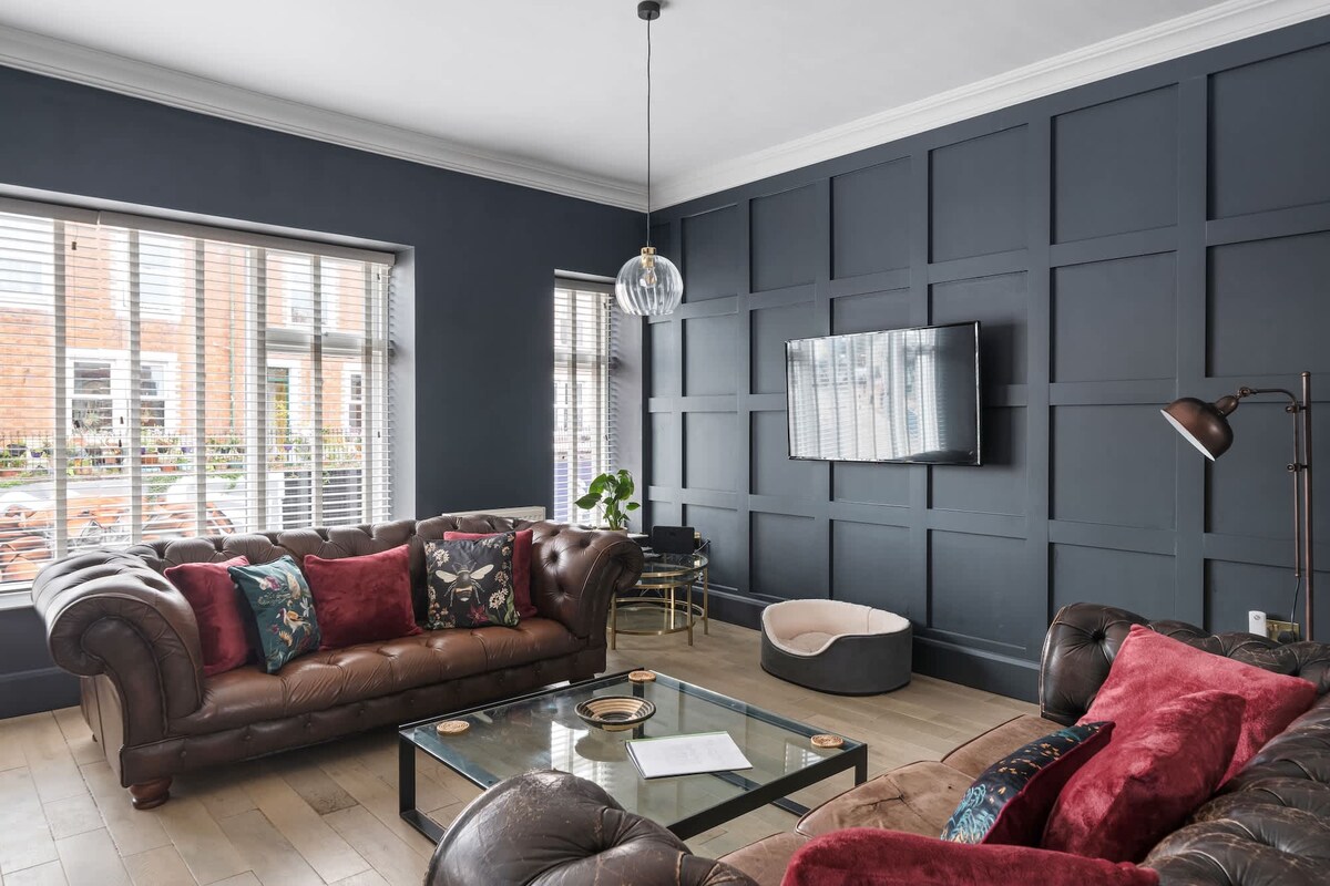 A stylish living area is highlighted by dark walls and natural light streaming through large windows. Two leather sofas with vibrant cushions surround a glass coffee table. A flat-screen television is mounted on the wall, while a cozy pet bed is positioned nearby.
