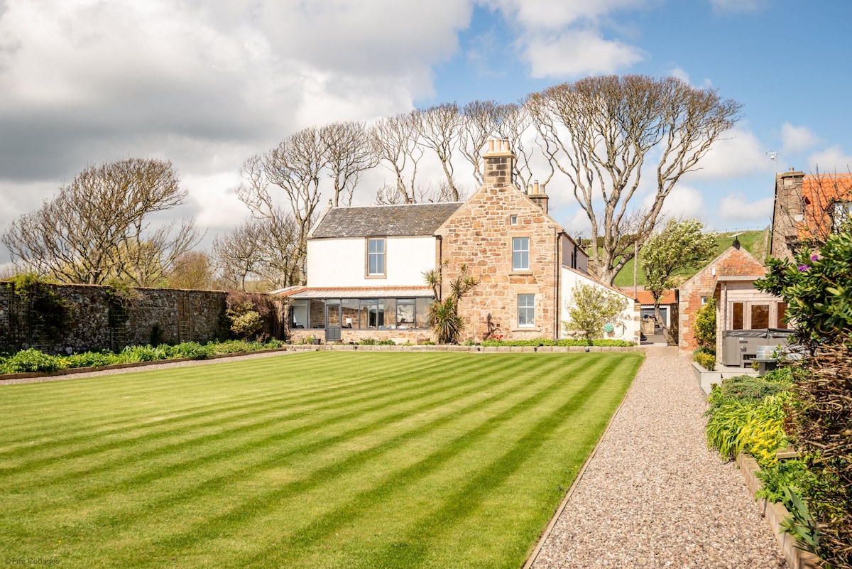 The exterior of Caiplie Farmhouse is featured, showcasing a well-maintained lawn bordered by manicured flower beds. The newly renovated farmhouse exhibits a blend of stone and modern architecture, with large windows and a covered patio area visible beneath the trees from the south-facing garden.