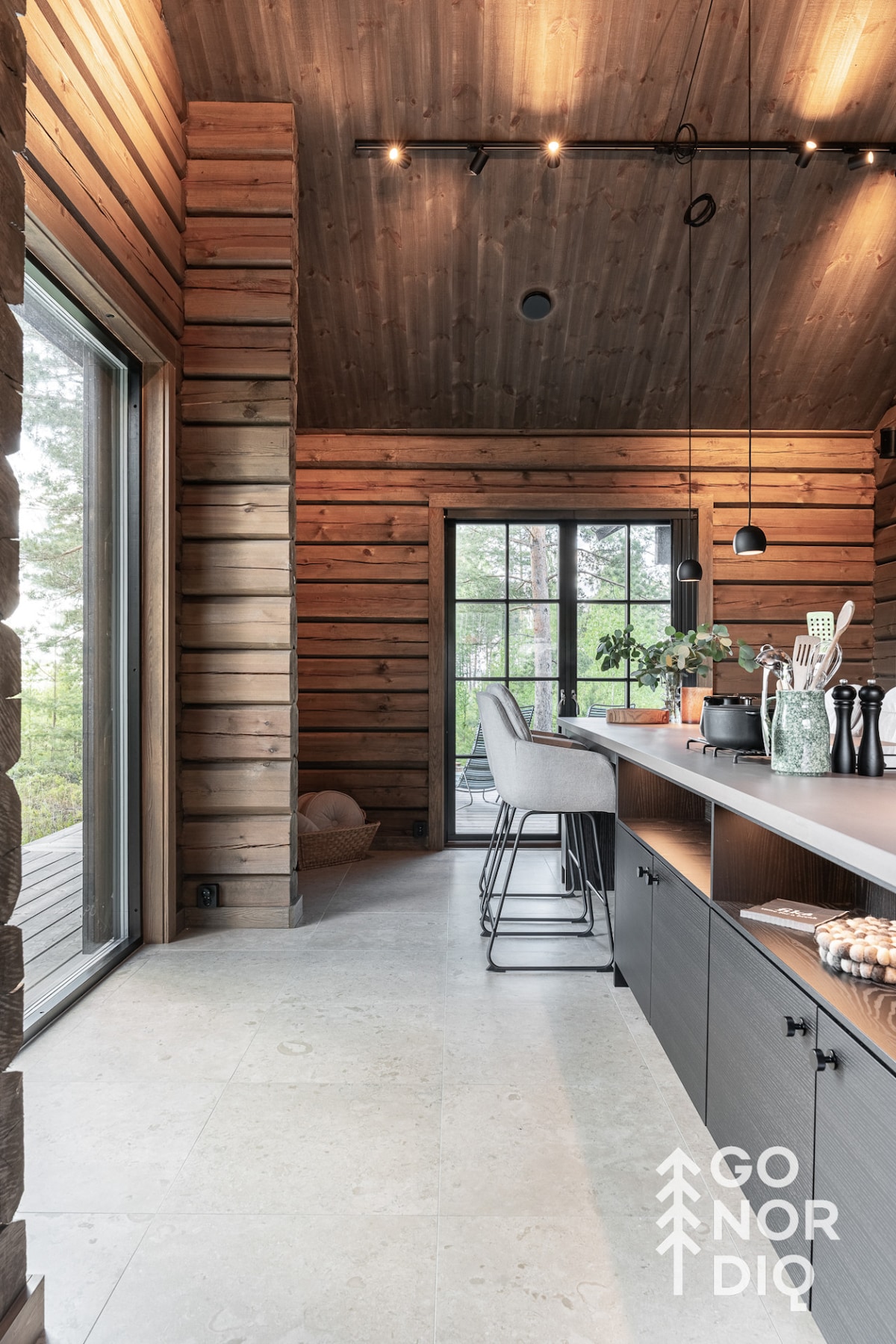 A spacious kitchen features a long countertop with modern appliances. A high-seated bar table encourages seating, complemented by sleek stools. Large windows invite natural light and offer views of the surrounding trees, enhancing the overall warm timber aesthetic.