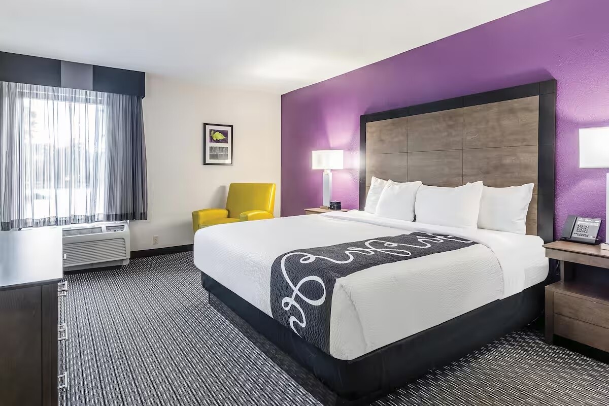 A spacious hotel room features a king bed with white linens against a purple accent wall. A cozy armchair in yellow adds a pop of color. Natural light enters through a large window, complemented by sheer and blackout curtains. A dresser and bedside tables are visible.