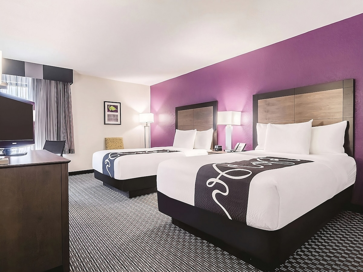 A room is furnished with two double beds, each with crisp white bedding. Soft purple walls complement the dark wood accents. A workspace desk and flat-screen TV are visible, while natural light streams through the window, adding brightness to the space.