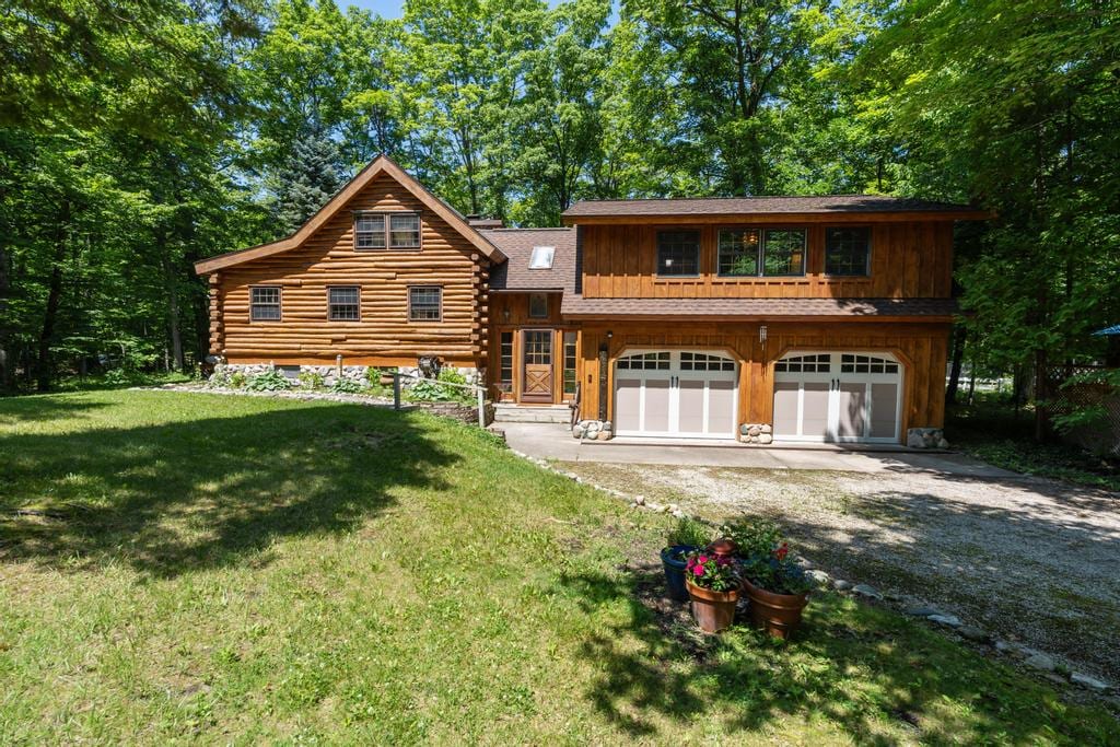 The exterior of the log cabin showcases a two-story structure surrounded by lush green trees. A well-maintained lawn features garden pots with colorful flowers. The entrance includes a wooden door and large windows, while a spacious driveway leads to the two-car garage.