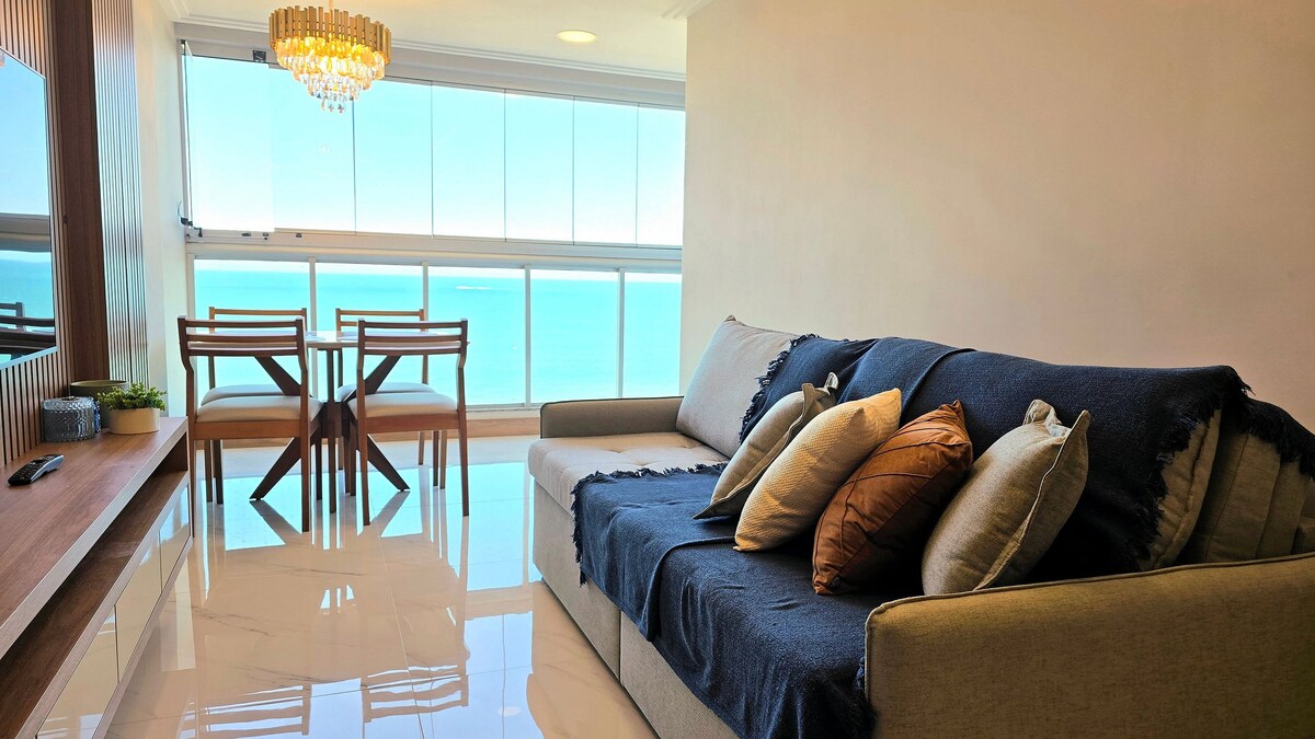 A bright living area is captured, featuring a comfortable sofa adorned with several decorative pillows. A dining table with four chairs is positioned near large glass windows, allowing for a view of the ocean. Natural light enhances the space.