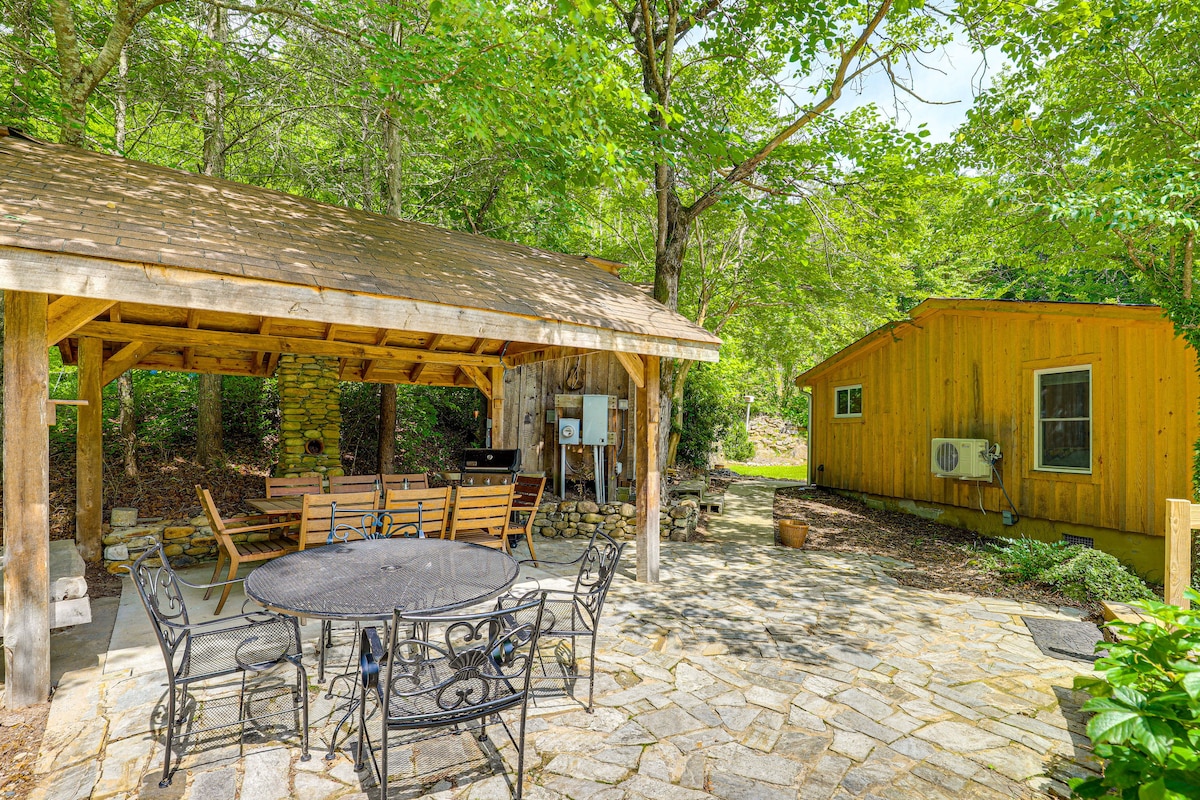 An outdoor patio area features a covered wooden structure with a built-in grill, complemented by a stone floor. A circular dining table with six chairs is arranged nearby, surrounded by lush greenery and trees, creating a tranquil space for gatherings.