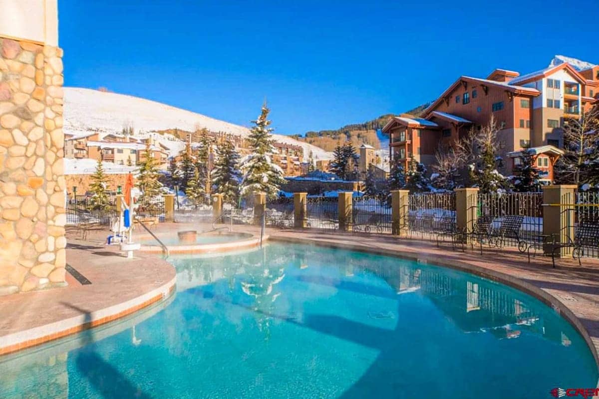 An outdoor pool is set against a backdrop of snow-covered mountains and trees. Steam rises gently from the water, and the pool area features a stone deck with lounge chairs and a nearby hot tub, creating a relaxing ambiance in a scenic environment.